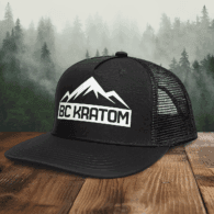 BC Kratom Trucker Snapback Hat placed on a wooden table in front of a scenic misty mountain landscape.