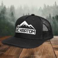 BC Kratom Trucker Snapback Hat placed on a wooden table in front of a scenic misty mountain landscape.
