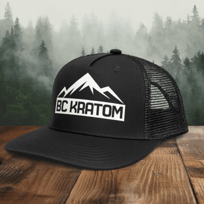 BC Kratom Trucker Snapback Hat placed on a wooden table in front of a scenic misty mountain landscape.
